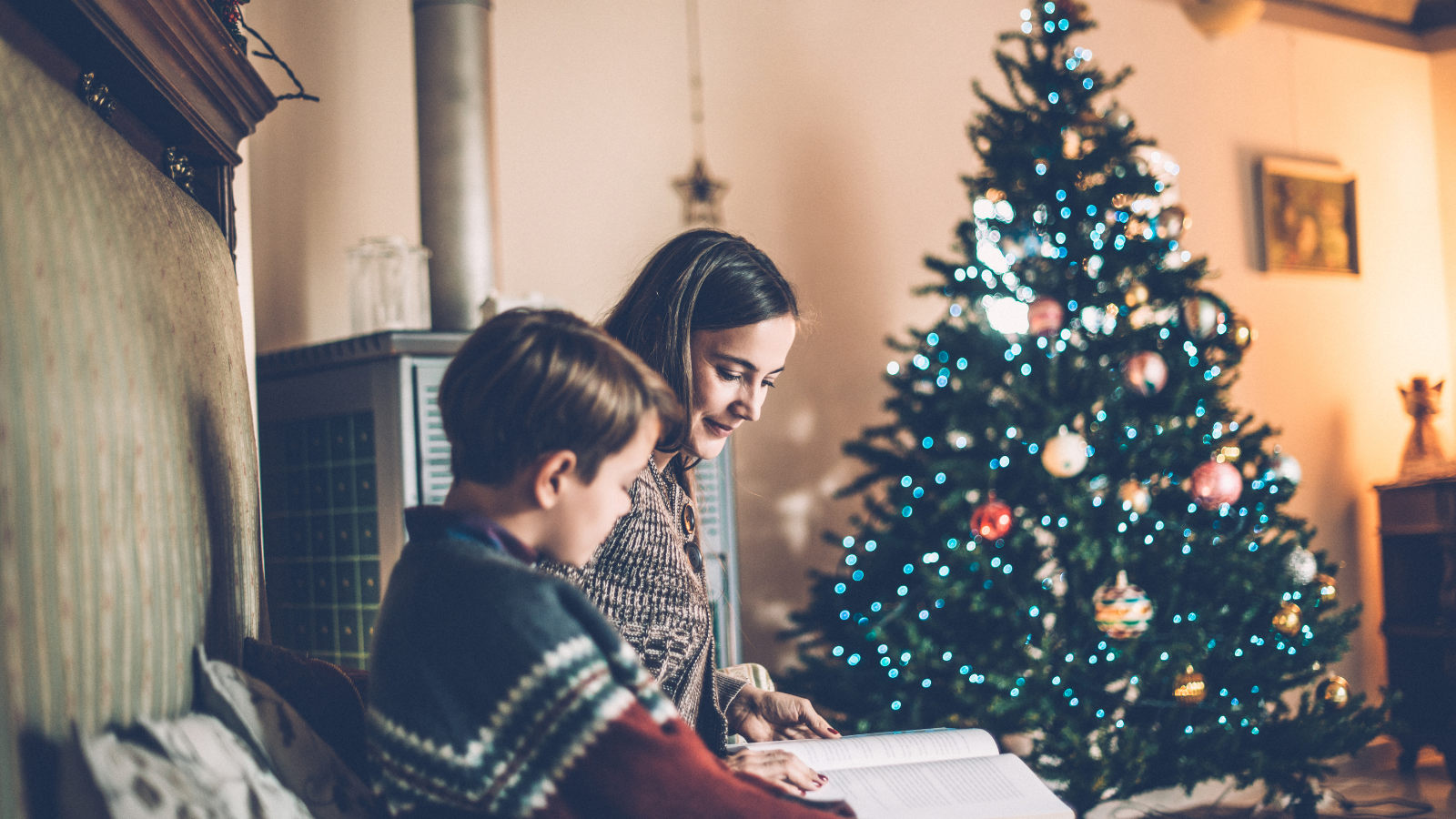 A woman reading a book to a boy with a Christmas tree in the background.